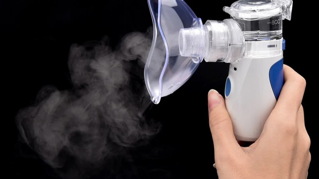 A close-up of a handheld nebulizer releasing mist, held by a person's hand against a black background.